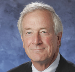 An older man with gray hair, likely among top Chicago lawyers, is wearing a dark suit and light blue shirt, posing confidently in front of a blue mottled background.