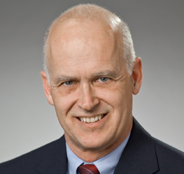 A middle-aged man with a bald head, light skin, and blue eyes, wearing a dark suit, light shirt, and red tie, smiles at the camera against a gray background—perfect for a corporate law office setting.