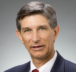 A middle-aged man in a dark suit, white shirt, and red tie poses for a professional headshot against a plain gray background, presenting the polished image expected at leading law offices.