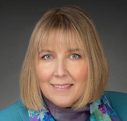 A woman with straight blonde hair and bangs, wearing a teal jacket and a colorful scarf, poses against a dark background—her confident look reminiscent of experienced lawyers in Chicago.