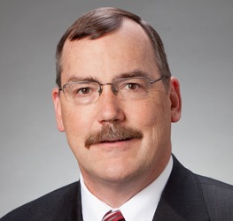A man with short brown hair, a mustache, and glasses, wearing a dark suit, white shirt, and striped tie, poses in front of a plain gray background—reflecting the professionalism of Chicago lawyers specializing in intellectual property law.
