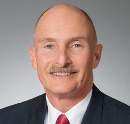A middle-aged man with a bald head and mustache, wearing a dark suit, white shirt, and red patterned tie, posed against a gray background—perfect for a corporate law office or litigation support professional.
