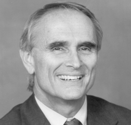 A grayscale portrait of an older man with light hair, wearing a suit and tie, smiling at the camera against a plain background, reflecting the professionalism often seen in corporate law office settings.