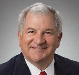 Smiling older man with short gray hair wearing a dark suit, white shirt, and red tie against a plain gray background, representing experienced lawyers in Chicago.