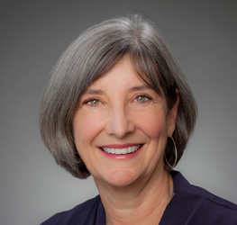 A woman with short gray hair and light eyes smiles at the camera. She is wearing a dark top and hoop earrings, with a neutral gray background—perfect for law offices or litigation support profiles.