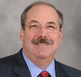Middle-aged man with glasses, a mustache, and short hair, wearing a suit, blue shirt, and red tie—posed in front of a gray background—ideal for a Chicago lawyers or corporate law office profile.