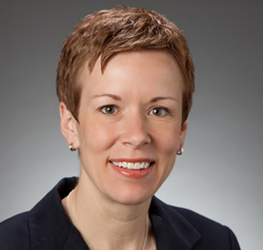 A woman with short light brown hair, wearing a dark blazer and a necklace, smiles at the camera against a plain gray background, reflecting the confidence and professionalism found in top Chicago lawyers.