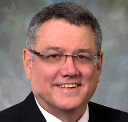 A middle-aged man with short gray hair, glasses, and a suit jacket smiles at the camera against a muted background, suggesting the professional atmosphere of a corporate law office.