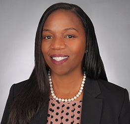 Woman with long straight hair wearing a black blazer, pink polka dot blouse, and pearl necklace, smiling confidently in front of a gray background—perfect for representing Chicago lawyers or law offices specializing in intellectual property law.