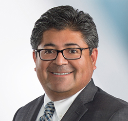 A man with short dark hair and glasses, wearing a dark suit, white shirt, and striped tie, smiles at the camera against a light background—representing chicago lawyers specializing in intellectual property law.