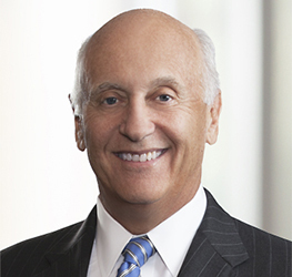 Smiling older man with short gray hair wearing a dark pinstripe suit, white shirt, and blue striped tie, posed against a light, blurred background—an experienced figure in Chicago lawyers and intellectual property law.