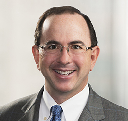A man with short brown hair, glasses, and a gray suit with a blue tie, smiling at the camera against a neutral blurred background, representing litigation support at a corporate law office.