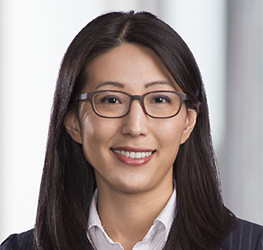 A woman with straight, dark hair and glasses, wearing a white shirt and dark blazer, smiles at the camera against a blurred background in a corporate law office.