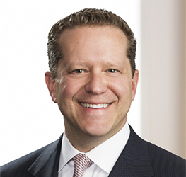 Smiling man in a dark suit, white shirt, and light-colored tie stands in front of a neutral background, reflecting the professionalism of leading law offices.