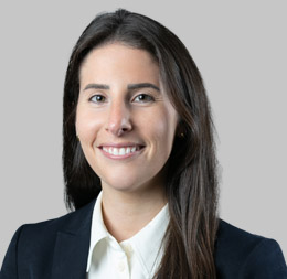 Woman with long dark hair wearing a black blazer and white shirt, smiling at the camera against a plain gray background, reflects professionalism in a corporate law office specializing in litigation support.