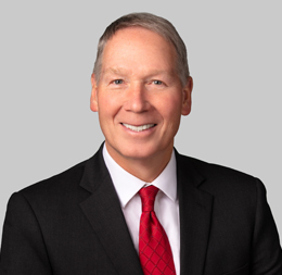 A man in a black suit, white shirt, and red tie is smiling at the camera against a plain light gray background, reflecting the professional image seen at top corporate law offices.