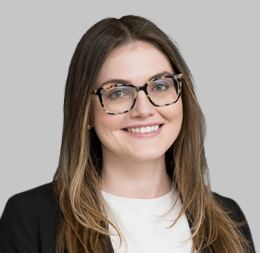 A woman with long brown hair, wearing glasses, a white top, and a black blazer, smiling in front of a plain gray background—perfectly reflecting the professional atmosphere of leading law offices in Chicago.