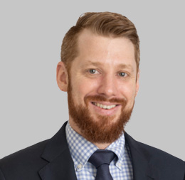 A man with light skin, short reddish-brown hair, and a trimmed beard, wearing a dark suit, blue checkered shirt, and navy tie, smiles at the camera against a gray background—representative of lawyers in Chicago corporate law offices.