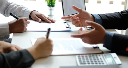 Three people sit at a table with documents, pens, a calculator, and a small house model, engaged in a discussion—perhaps consulting lawyers in Chicago about real estate or intellectual property law.