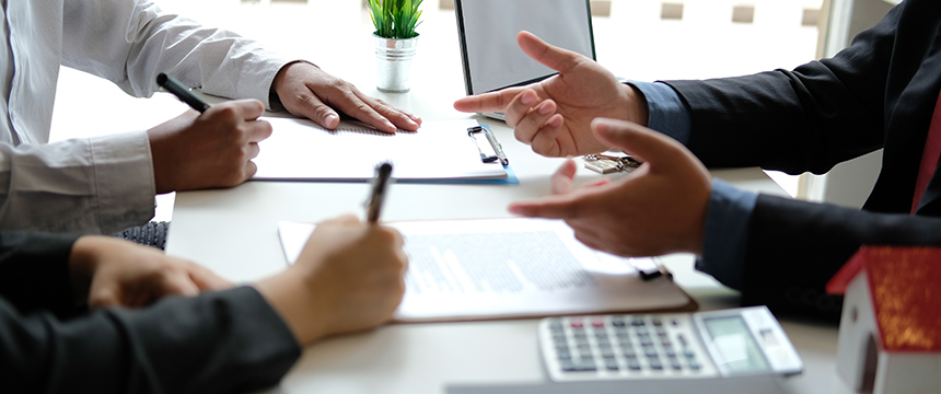 Three people sit at a table with documents, pens, a calculator, and a small house model, engaged in a discussion—perhaps consulting lawyers in Chicago about real estate or intellectual property law.