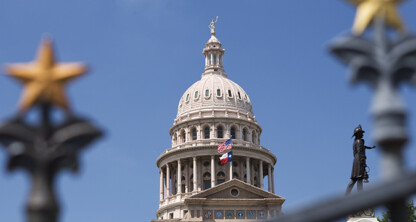The Texas State Capitol dome is seen through a fence with gold stars, under a clear blue sky, with a statue visible on the right—an inspiring view for any corporate law office or those specializing in intellectual property law.