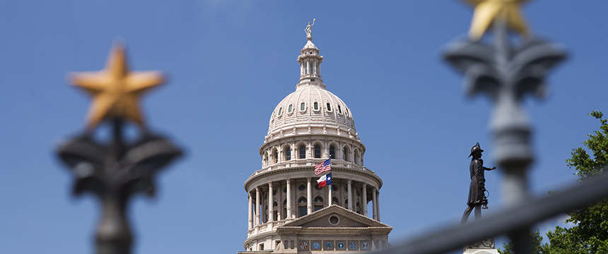 The Texas State Capitol dome is seen through a fence with gold stars, under a clear blue sky, with a statue visible on the right—an inspiring view for any corporate law office or those specializing in intellectual property law.