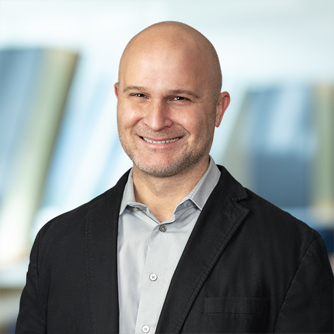 A bald man in a light gray shirt and dark blazer smiles in front of a blurred background with blue and beige tones, representing professionals who offer litigation support and intellectual property law services.