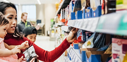 A woman holds a toddler while browsing products on a supermarket shelf with a young child beside her, perhaps picking up snacks after visiting lawyers in Chicago.