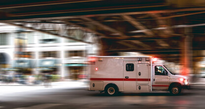 An ambulance with flashing lights drives quickly under an overpass in an urban area, passing by law offices offering litigation support.