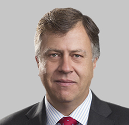 A middle-aged man with short brown hair, wearing a dark suit, light blue striped shirt, and red tie, pictured against a plain light gray background, embodies the professionalism of a corporate law office.