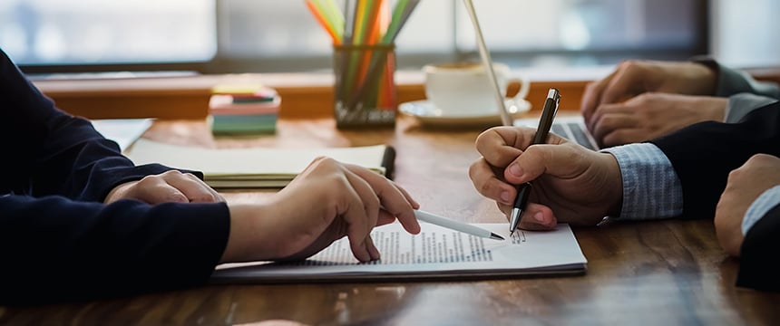 Two people sit at a wooden desk reviewing and signing documents with pens, while Chicago lawyers provide guidance. Office supplies and a cup appear in the background.