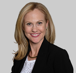 A woman with long blonde hair is wearing a black blazer over a white top and smiling at the camera against a plain gray background, presenting a professional image typical of a corporate law office.