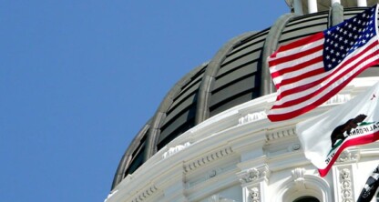 The U.S. and California state flags fly in front of the dome of a government building against a clear blue sky, symbolizing justice and governance often represented by nearby law offices or litigation support services.