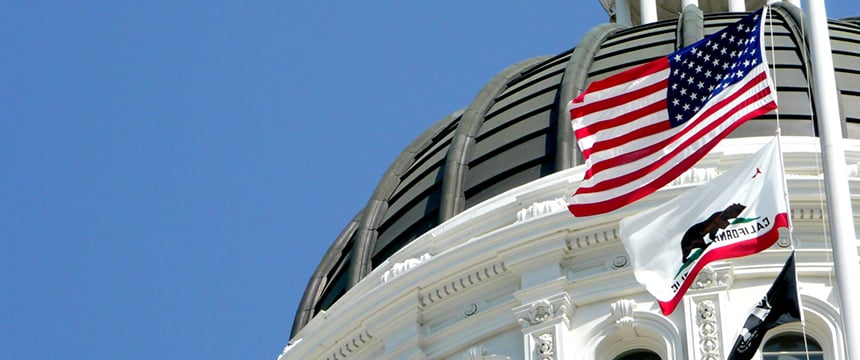 The U.S. and California state flags fly in front of the dome of a government building against a clear blue sky, symbolizing justice and governance often represented by nearby law offices or litigation support services.