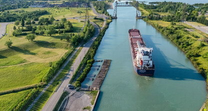 A large cargo ship travels along a narrow canal bordered by green fields and trees, with a bridge visible in the distance under a clear sky—reminiscent of the precision and strategy found in a corporate law office.