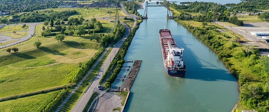 A large cargo ship travels along a narrow canal bordered by green fields and trees, with a bridge visible in the distance under a clear sky—reminiscent of the precision and strategy found in a corporate law office.
