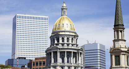A view of the Colorado State Capitol with its gold dome, surrounded by modern office buildings, a corporate law office, and a church steeple, under a partly cloudy sky.