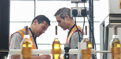 Two workers in safety vests inspect equipment in a factory with bottles on a conveyor belt; a monitor and control panel are visible, highlighting the importance of protecting innovations with intellectual property law.