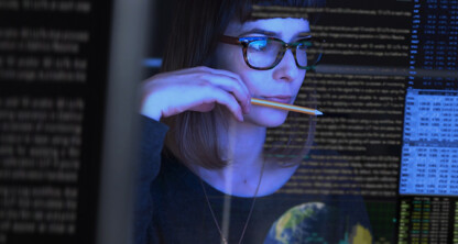 A woman with glasses looks at multiple computer screens displaying lines of code and data, holding a pencil to her mouth in concentration, as she researches intellectual property law for chicago lawyers.