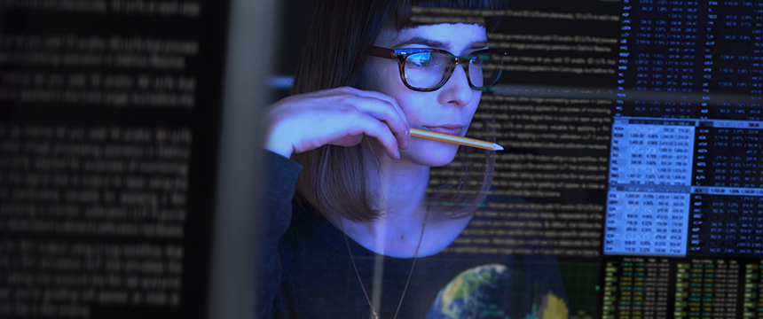 A woman with glasses looks at multiple computer screens displaying lines of code and data, holding a pencil to her mouth in concentration, as she researches intellectual property law for chicago lawyers.