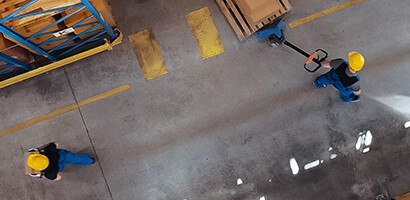 Warehouse interior viewed from above showing two workers in blue uniforms and yellow helmets, one pushing a hand pallet truck among shelves of boxes and pallets—ideal for businesses seeking lawyers in Chicago with expertise in intellectual property law.