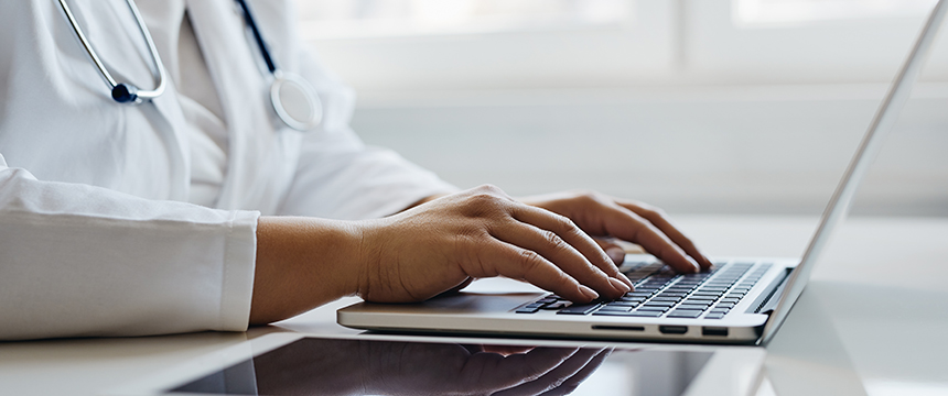 A person in a white coat with a stethoscope types on a laptop at a desk, supporting Chicago lawyers and law offices with litigation support, while a tablet lies next to the computer.