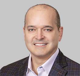 A middle-aged man with light skin, a shaved head, and a checked blazer over a light-colored shirt smiles at the camera against a plain gray background, reflecting the professionalism of lawyers in Chicago specializing in intellectual property law.