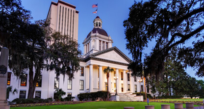 The image shows the Florida State Capitol building in Tallahassee at dusk, featuring white columns, a central dome, and a tall office tower—an impressive setting reminiscent of prestigious law offices or litigation support centers.
