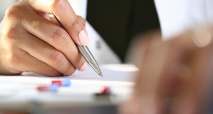 A person holding a pen writes on paper near assorted pills on a desk, with another hand and blurred foreground visible—evoking the careful documentation often seen in corporate law office settings.