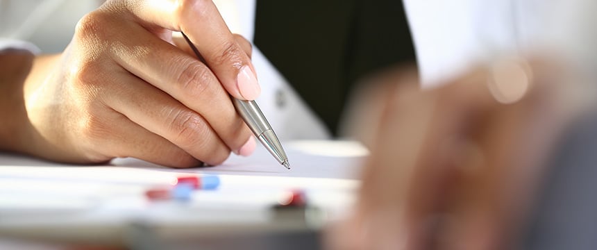 A person holding a pen writes on paper near assorted pills on a desk, with another hand and blurred foreground visible—evoking the careful documentation often seen in corporate law office settings.