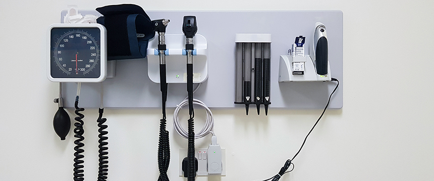 Wall-mounted medical equipment including a blood pressure monitor, otoscope, ophthalmoscope, thermometer, and containers for disposable covers in a clinical setting often found in Chicago lawyers’ offices offering on-site health services.