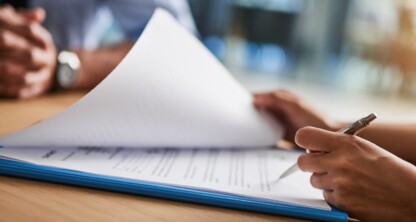 A person holding a pen reviews and flips through documents on a clipboard at a table in a corporate law office, with another individual seated across from them.
