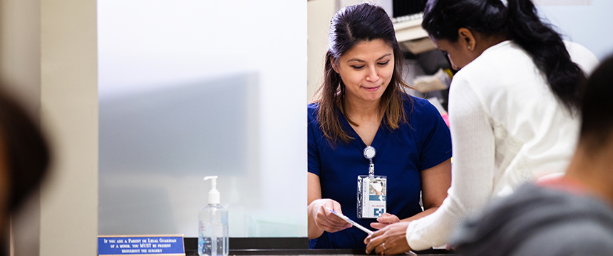 A woman in blue scrubs assists another person at a reception desk, handing over a card. A bottle of hand sanitizer is on the counter, highlighting the attention to detail often valued by lawyers in Chicago seeking reliable litigation support.