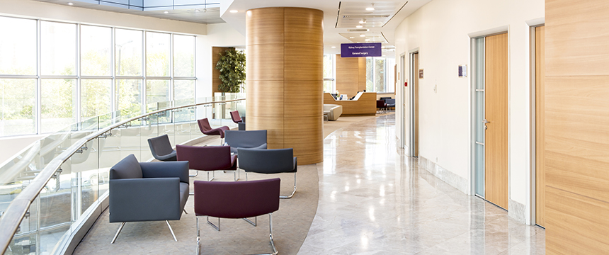 Modern hospital waiting area with grey and purple chairs, a curved glass railing, wood-paneled columns, and large windows letting in natural light—ideal for consultations or meetings with lawyers in Chicago. Reception desk visible in the background.